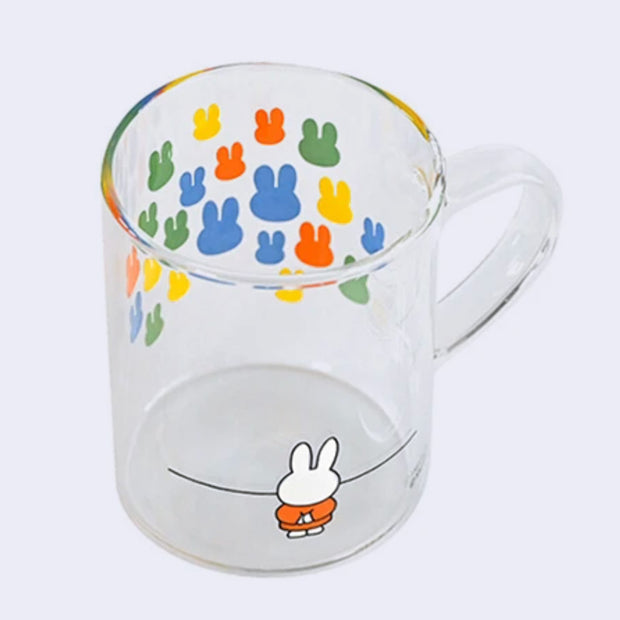 Glass mug with a wrap around graphic of Miffy, standing with her arms behind her back and looking at a large colorful array of solid colored bunny heads on a wall, as though in a museum.