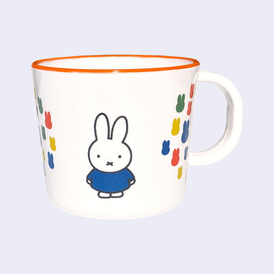 White mug with an orange rim of Miffy, standing with her arms at her sides and wearing a blue dress. Graphics on other side of the cup show several primary colored silhouettes of Miffy's head, arranged together closely like a collage.