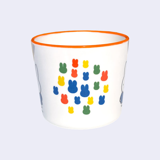 White mug with an orange rim of Miffy, standing with her arms at her sides and wearing a blue dress. Graphics on other side of the cup show several primary colored silhouettes of Miffy's head, arranged together closely like a collage.