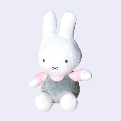 Plush doll of a white bunny with a large head and arms outspread. It wears a light. pink shirt and gray overalls and sits.