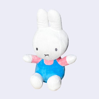 Plush doll of a white bunny with a large head and arms outspread. It wears a bright pink shirt and blue overalls and sits.