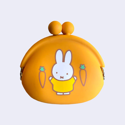 Orange silicone coin pouch with an illustration of Miffy wearing a yellow dress and standing with her arms up. Besides her are 2 carrots with short green stems.