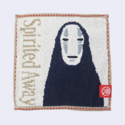 Small square cream colored towel with a tan outline and sideways text that reads "Spirited Away." The character No Face is next to the text, seen from the mid section up.