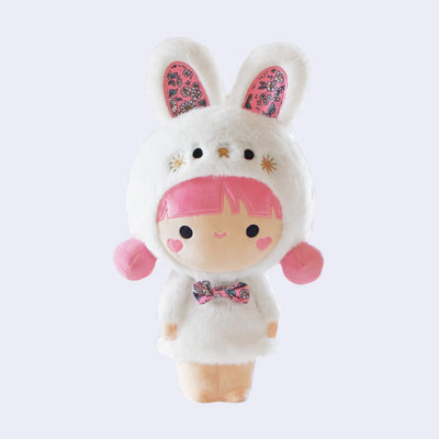 Plush doll of a girl with pink hair and rosy red cheeks. She smiles and is dressed in a fluffy white dress with matching bunny shaped hood hat with ears. She has a floral pattern bowtie and floral pattern inside the bunny ears.