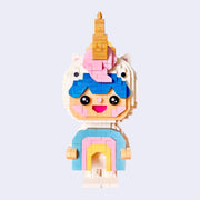 3D sculpture made out of small brick like plastic pieces of a blue haired boy wearing a white body suit and a unicorn hood hat, with pink mane and golden horn. He smiles and holds a pastel colored rainbow.