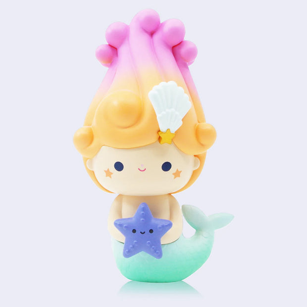 Vinyl figure of a girl with a teal blue mermaid tail and a swirl of blonde to pink gradient hair atop her head in a semi cone pattern.  She has a cute smile and holds a purple star fish in her hands.