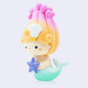 Vinyl figure of a girl with a teal blue mermaid tail and a swirl of blonde to pink gradient hair atop her head in a semi cone pattern.  She has a cute smile and holds a purple star fish in her hands.