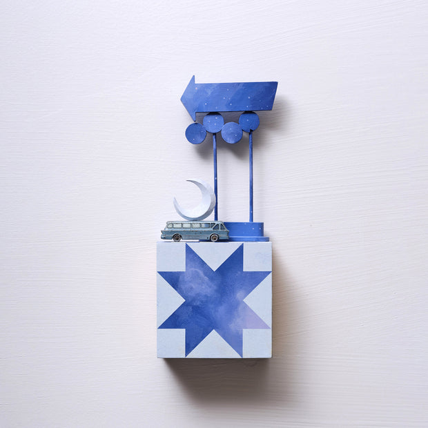 Small square wood panel painted like a dark blue sky with light blue geometric patterning overlayed on top, making a star shaped tile design. Atop of it is a small model bus and wooden sculptures of a crescent moon and a series of large Americana style signs attached to the same poles.