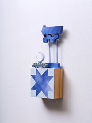 Small square wood panel painted like a dark blue sky with light blue geometric patterning overlayed on top, making a star shaped tile design. Atop of it is a small model bus and wooden sculptures of a crescent moon and a series of large Americana style signs attached to the same poles.