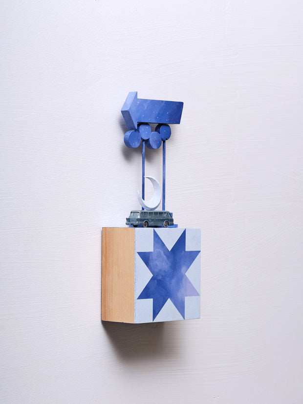 Small square wood panel painted like a dark blue sky with light blue geometric patterning overlayed on top, making a star shaped tile design. Atop of it is a small model bus and wooden sculptures of a crescent moon and a series of large Americana style signs attached to the same poles.