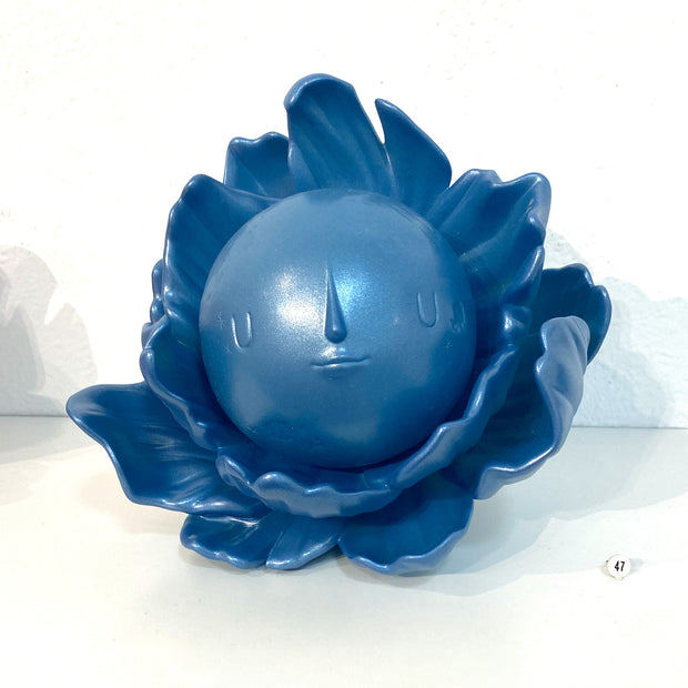 Steel blue colored vinyl figure with a glittery surface shimmer of a round full moon with a calm, minimalistic face. It sits within a series of flower petals, as if it is the pistil of the flower.