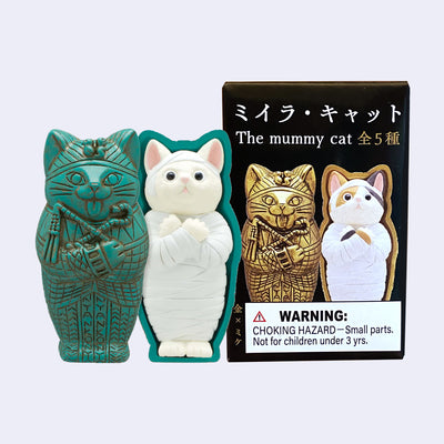 Small plastic figurine of a green oxidized sarcophagus shaped like a cat with a white mummified cat inside. It stands next to its product packaging.