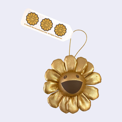 A small plush Murakami flower with a cheerful expression made out of a gold fabric, with a hanging tag attached.