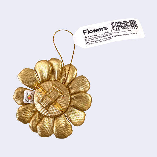 A small plush Murakami flower made from a shiny gold fabric with a brooch pin backing attached to it and a hanging string.