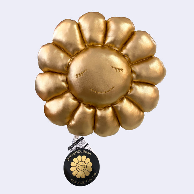 A plush Murakami flower with a calm, closed eye but smiling expression made out of a gold fabric with tags still attached.