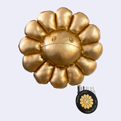 A plush Murakami flower with a cheerful expression made out of a gold fabric with tags still attached.