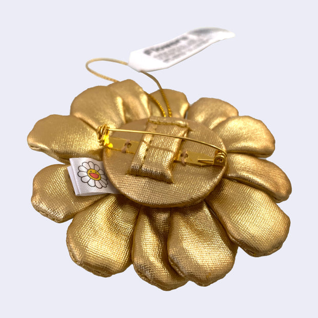 A small plush Murakami flower made from a shiny gold fabric with a brooch pin backing attached to it and a hanging string.