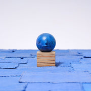 Small carved wooden sculpture of a dark blue moon with a closed eye, calm smiling expression. It rests atop of a striated grain wooden square pedestal. 