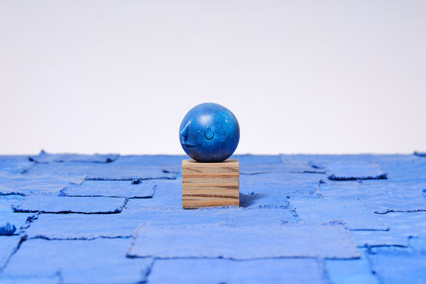 Small carved wooden sculpture of a dark blue moon with a closed eye, calm smiling expression. It rests atop of a striated grain wooden square pedestal. 