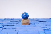 Small carved wooden sculpture of a dark blue moon with a closed eye, calm smiling expression. It rests atop of a striated grain wooden square pedestal. 