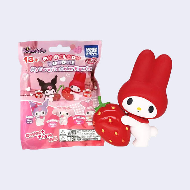 Figure of My Melody wearing a red hood and holding onto a large strawberry, with a surprised expression. She stands next to her product packaging, a bag with photos of all the mystery options inside.