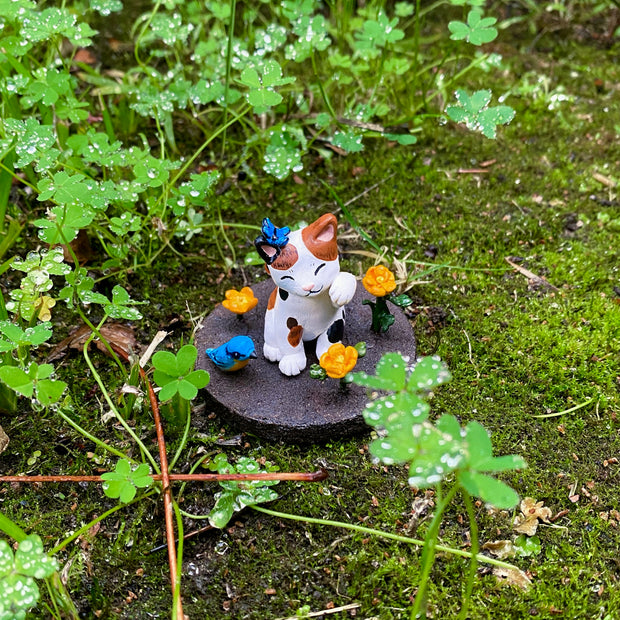 Sculpture of a calico cat sitting with one paw raised and a closed eye, smiling expression. A blue butterfly sits atop its head. The cat holds an orange flower in its tail and sits on a brown dirt circular base with orange flowers and a blue and orange bird.