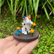 Sculpture of a calico cat sitting with one paw raised and a closed eye, smiling expression. A blue butterfly sits atop its head. The cat sits on a brown dirt circular base with orange flowers and a blue and orange bird.