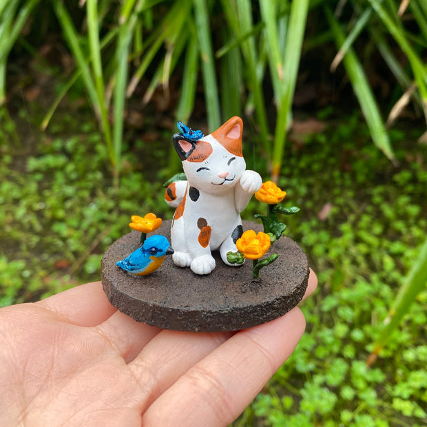 Sculpture of a calico cat sitting with one paw raised and a closed eye, smiling expression. A blue butterfly sits atop its head. The cat sits on a brown dirt circular base with orange flowers and a blue and orange bird.