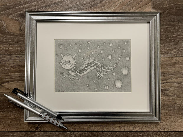 Graphite drawing of a cartoon style dragon, with many scales and floating lanterns in the background. Piece is in silver frame.