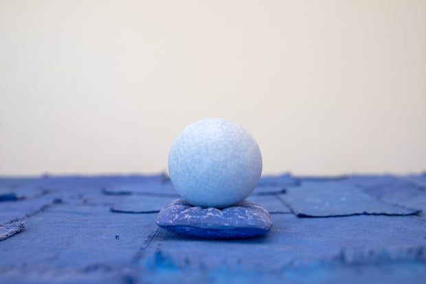 Small carved wooden sculpture of a blue sphere with a closed eye, calm smiling expression. It rests atop of a purple wooden pillow with splatter pattern, carved to look soft.