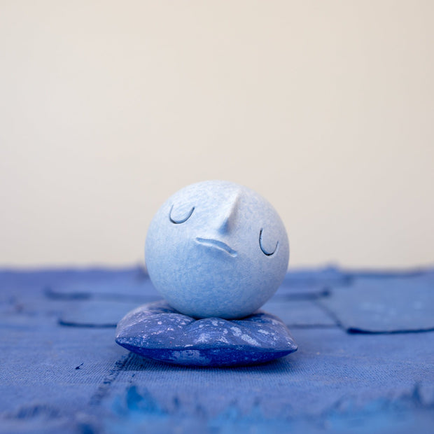 Small carved wooden sculpture of a blue sphere with a closed eye, calm smiling expression. It rests atop of a purple wooden pillow with splatter pattern, carved to look soft.