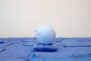 Small carved wooden sculpture of a blue sphere with a closed eye, calm smiling expression. It rests atop of a purple wooden pillow with splatter pattern, carved to look soft.