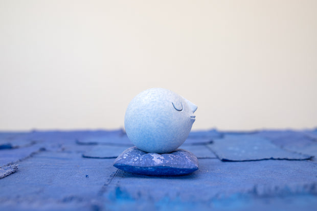 Small carved wooden sculpture of a blue sphere with a closed eye, calm smiling expression. It rests atop of a purple wooden pillow with splatter pattern, carved to look soft.