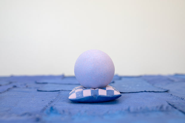 Small carved wooden sculpture of a purple sphere with a closed eye, calm smiling expression. It rests atop of a purple and pink grid pattern wooden pillow, carved to look soft.