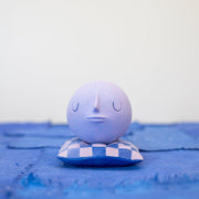 Small carved wooden sculpture of a purple sphere with a closed eye, calm smiling expression. It rests atop of a purple and pink grid pattern wooden pillow, carved to look soft.