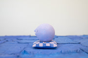 Small carved wooden sculpture of a purple sphere with a closed eye, calm smiling expression. It rests atop of a purple and pink grid pattern wooden pillow, carved to look soft.