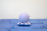 Small carved wooden sculpture of a purple sphere with a closed eye, calm smiling expression. It rests atop of a purple and pink grid pattern wooden pillow, carved to look soft.