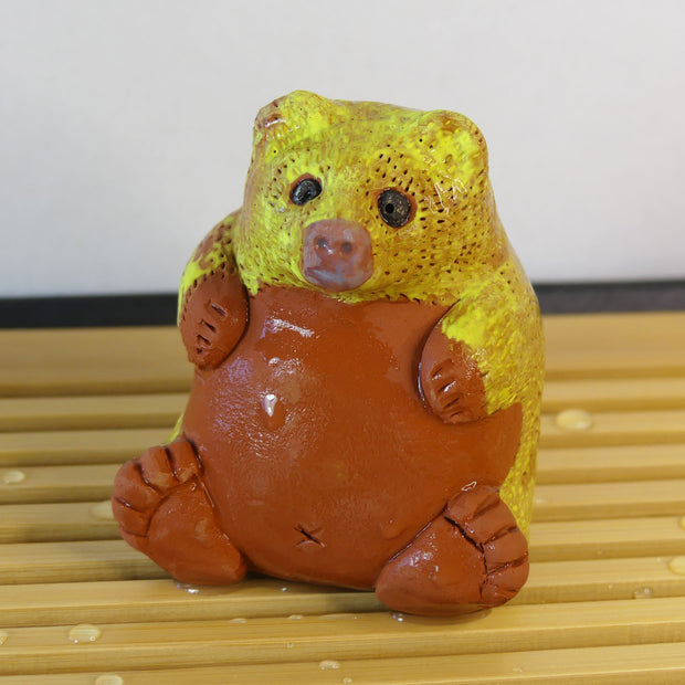 Small sculpture of a brown bear, chubby and sitting on its bottom. It has a yellow coloring for the top half of its body.