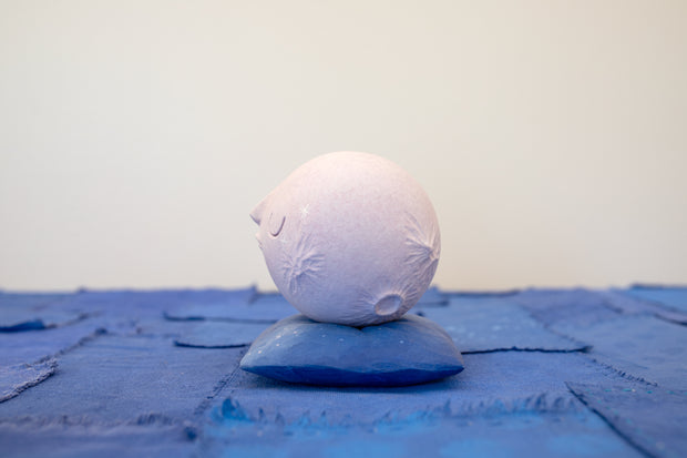 Small carved wooden sculpture of a light purple moon with a closed eye, calm smiling expression. It rests atop of a purple wooden pillow with splatter pattern, carved to look soft.