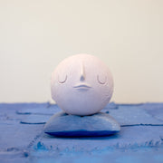 Small carved wooden sculpture of a light purple moon with a closed eye, calm smiling expression. It rests atop of a purple wooden pillow with splatter pattern, carved to look soft.