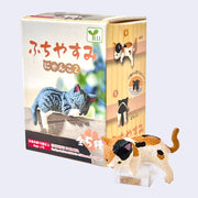 Small figure of a plastic calico cat, resting atop of a clear block with its product packaging behind.