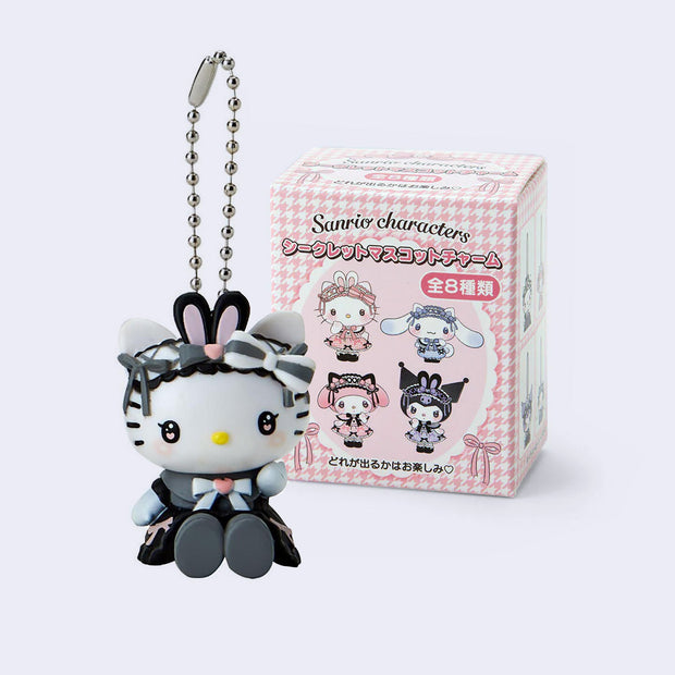 Figure of Hello Kitty dressed in a gothic Lolita style dress with dreamy eyes and blushed cheeks. She is attached to a ball chain and sits in front of her product packaging.