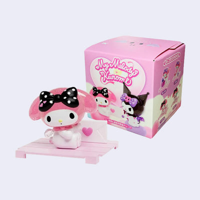 Vinyl figure of My Melody wearing a black and white polka dot bowtie, sitting on a pink wooden platform and holding an envelope, sealed with a heart sticker. She sits in front of her product packaging.