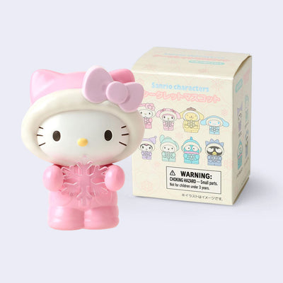 Figurines of Hello Kitty, dressed in a fluffy pink snowsuit with a fur lined hood pulled on. She still has her iconic bow, pink over her hood and holds a snowflake. She stands in front of product packaging.