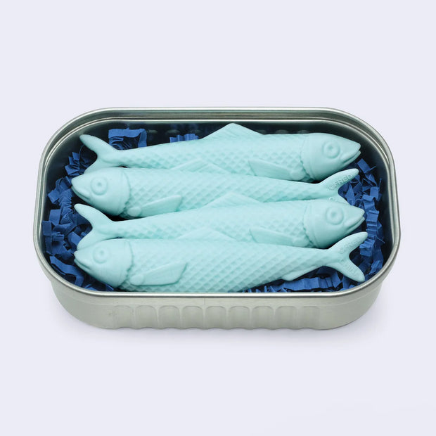 4 pale blue erasers shaped like sardines, packed together closely inside of a silver tin with blue scrunched paper.