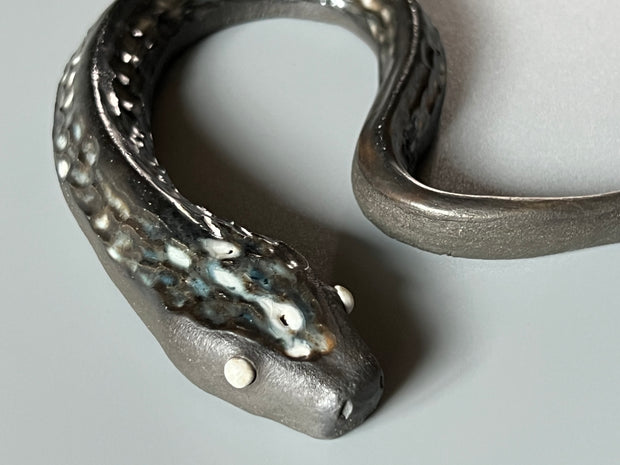 Ceramic sculpture of a dark gray snake with light silver eyes and imprinted scales along its body. Its tail curves slightly inward towards its head.