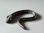 Ceramic sculpture of a dark gray snake with light silver eyes and imprinted scales along its body. Its tail curves slightly inward towards its head.