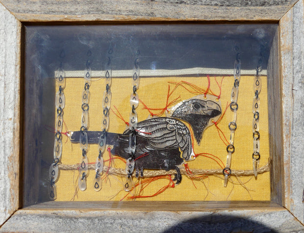 Mixed media sculpture in a wooden shadowbox frame of a bird sitting on a piece of twine with several red threads coming out from the bird. Rows of small plastic drawn rain drops hang along the top of the piece.