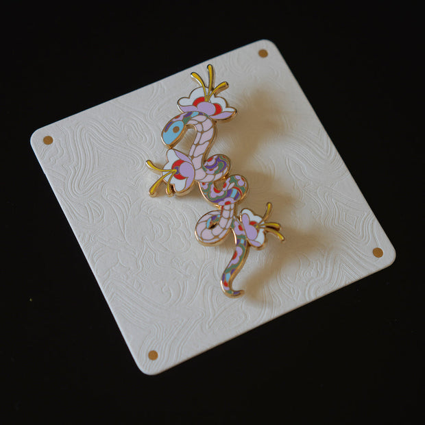 Enamel pin with gold outlining of a pastel colored snake, with its body elongated with small twists. The pattern of its scales is floral in nature and 3 flowers with blooming from the snake's body. Seen at an angle to show sheen.