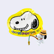 Small figure of Snoopy wearing a brown hat and sitting, holding a fishing rod. He sits in front of a yellow blind bag with Snoopy's smiling face on it.